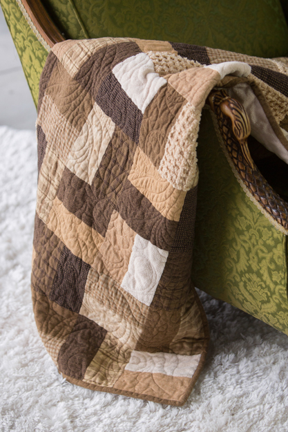Neutral brown patchwork quilt draped over a chair, showing classic tones and stitched quilting lines.
