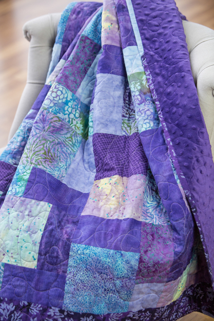 Purple patchwork quilt draped over a chair, showing rich colors and quilting texture.
